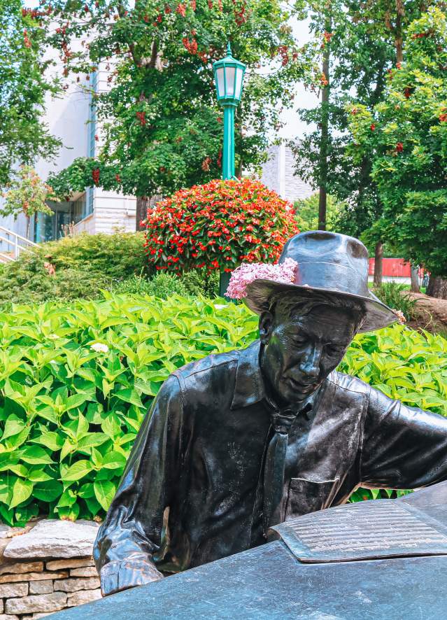 Hoagy Carmichael statue at Indiana University in Bloomington, Indiana, featuring a bronze figure seated at a piano in a landscaped garden with trees, shrubs, and a walkway in the background.