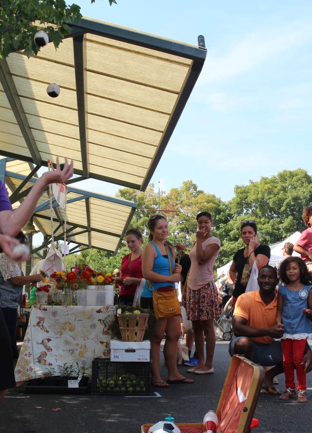 A man juggling balls at a Farmers Market in Bloomington, IN, while a diverse group of people watches under canopy tents on a sunny day.