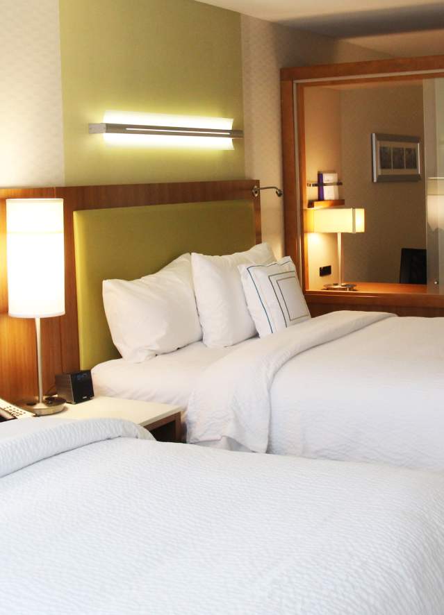 SpringHill Suites Hotel room interior with two neatly made beds, modern lighting, a bedside table, and a seating area with a sofa and lamp in the background.