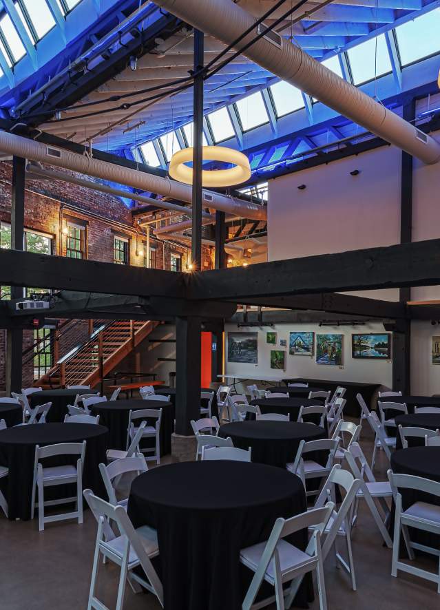 Bloomington event space with round tables covered in black cloths, white chairs, exposed brick walls, large windows, and circular ceiling lights.