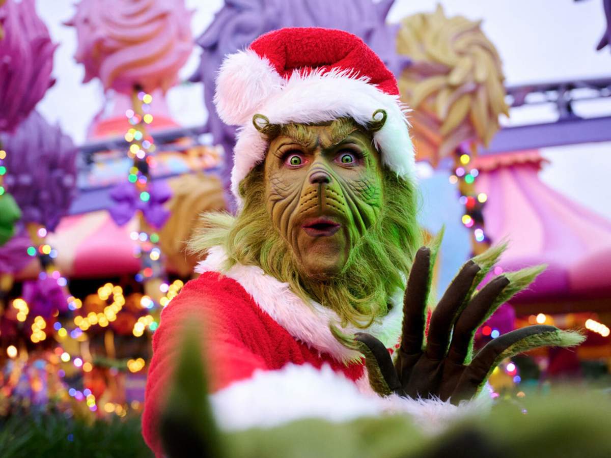 The Grinch reaches for the camera with the colorful holiday lights of Seuss Landing in the background