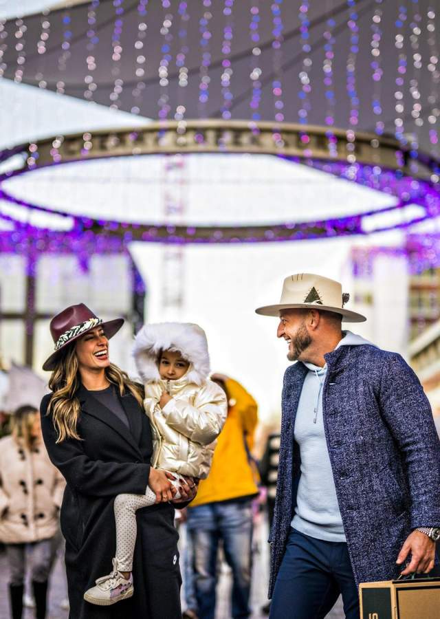 A family smiles as they walk through a holiday market in Denver's Cherry Creek neighborhood surrounded by colorful lights