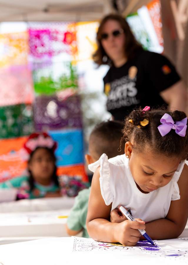 Young children coloring at an outdoor community event with colorful papel picado banners in the background.