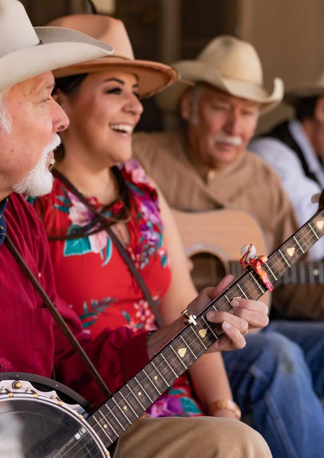 An older man with facial hair sits in the front playing a banjo. He his wearing a red button up shirt, a blue bandana, and a tan cowboy hat. A woman in red, a man playing a guitar, and a man playing harmonica are sitting behind him.