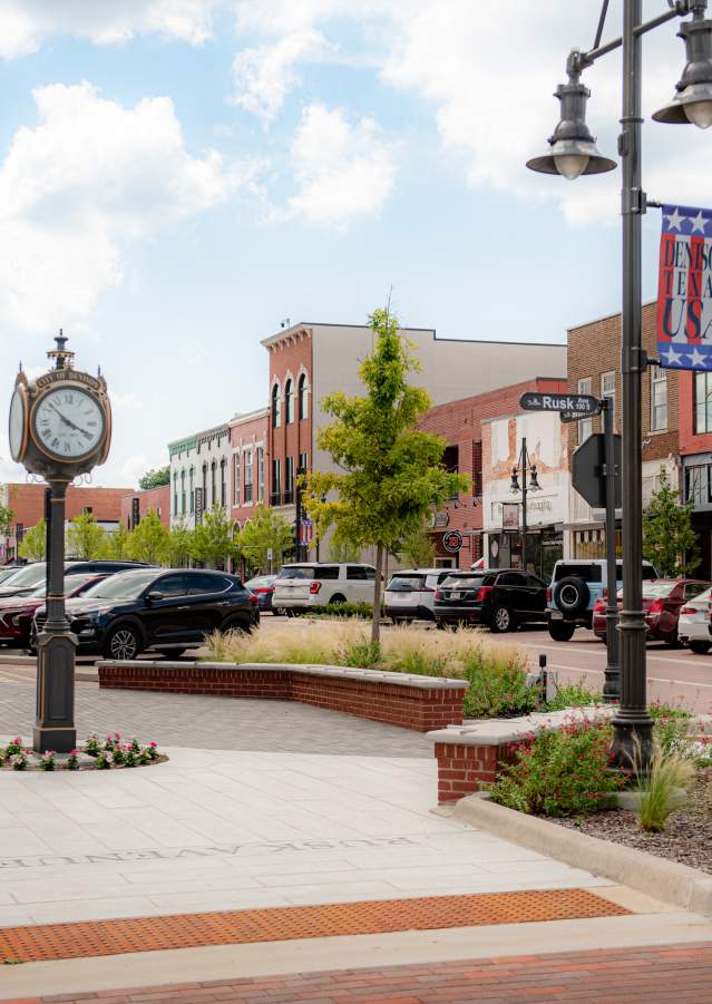 Picture of the 300 Block in Downtown Denison, Texas. Showcases the clock in front of City  Hall, the historic buildings in the background, cars of the visitors inside the shops that line up the Main Street