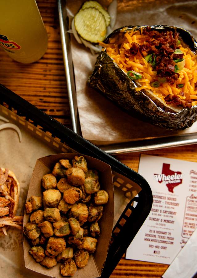 Two trays of food from Wheeler Barbeque viewed from above. One tray has a pulled pork sandwich with the top bun set to the side, a side of fried okra, and a couple of sliced pickles on the side. The other tray shows a baked potato loaded with cheese, green onions, and bacon with a slice of bread on the side.