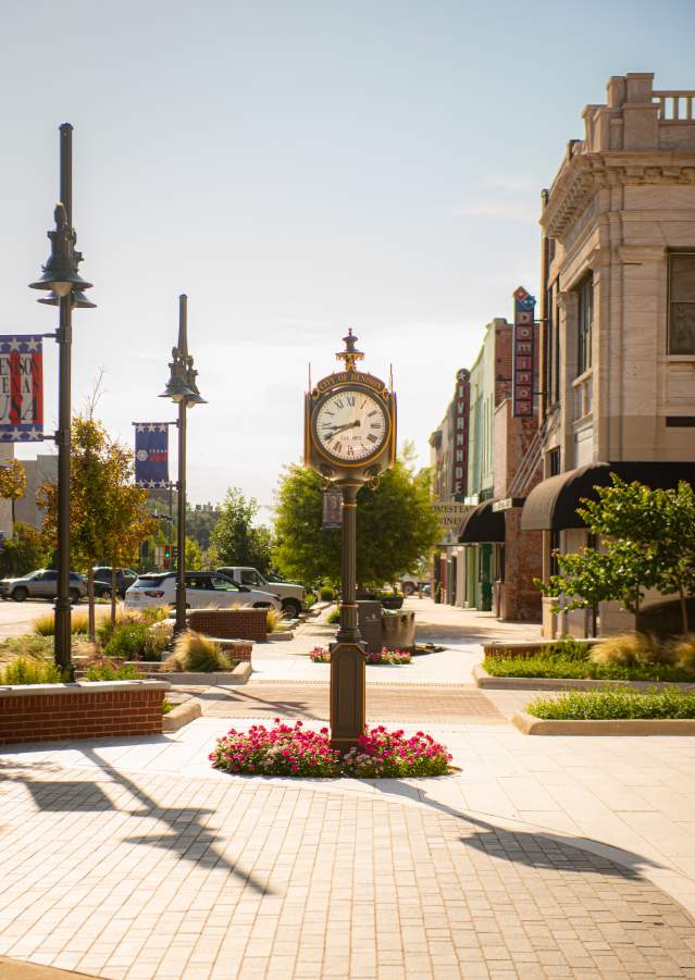 The City of Denison clock that is located in front of City Hall. It is a bright sunny day with clear blue sky. Pink flowers are in the bed around the base of the clock.