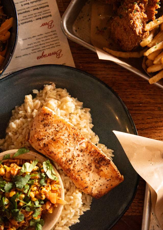 A top down view of a table filled with food at the George Tavern. The central plate has a piece of baked salmon on a bed of rice with a side bowl of Mexican street style corn. The bowl in the upper left corner is filled with a pasta dish. The upper right tray contains some chicken tenders and a pile of French fries.