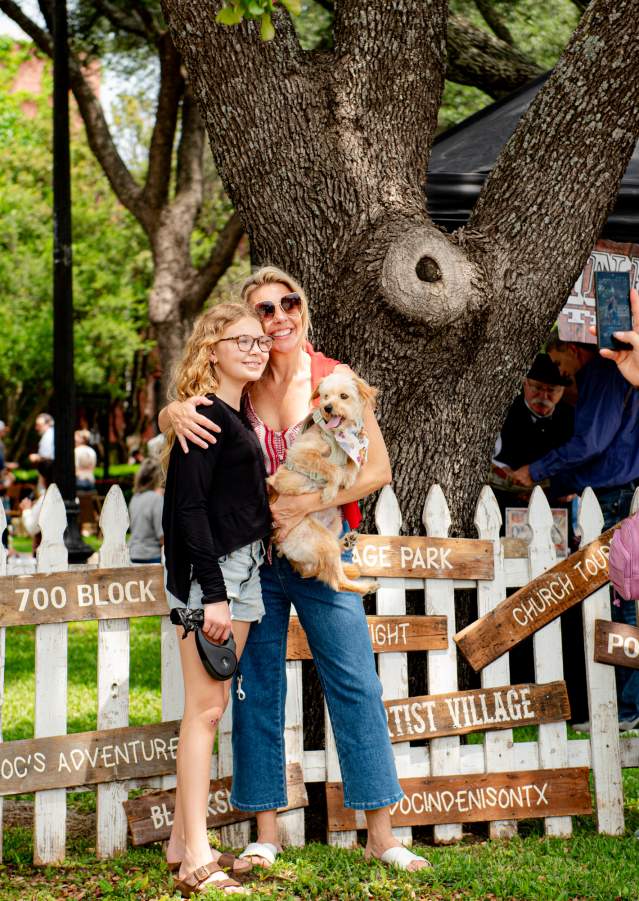 A blonde middle aged woman stands with one arm holding a small dog and the other arm draped over the solder of a younger girl with blonde curly hair. They are smiling and standing in front of a white picket fence decoration at the Doc Holliday Festival and a man in a white shirt is taking a picture of them with a cell phone.