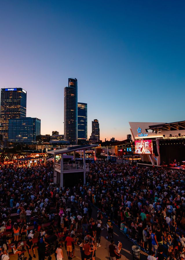 A large crowd gathers at the US Cellular Connection Stage during Summerfest in Milwaukee as the sun sets behind the city skyline, with high-rise buildings including the Baird Center visible in the background.