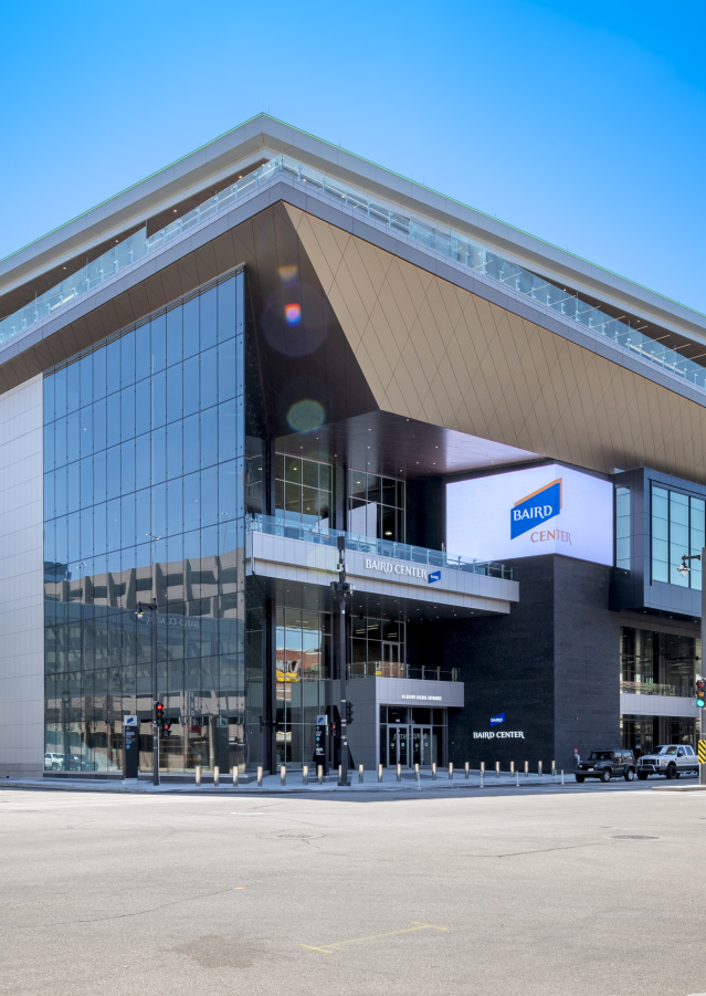 A daytime exterior view of the Baird Center in Milwaukee, showing its modern glass façade, angular roofline, and skywalk connection over the street, with cars and pedestrians nearby.