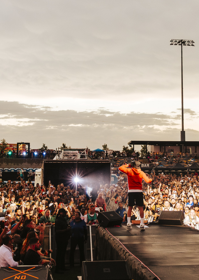 A performer in a red hoodie stands on an outdoor stage facing a massive crowd at sunset. Thousands of fans pack the venue, many holding up phones to capture the moment, while bright stadium lights and food stand signs glow in the background.