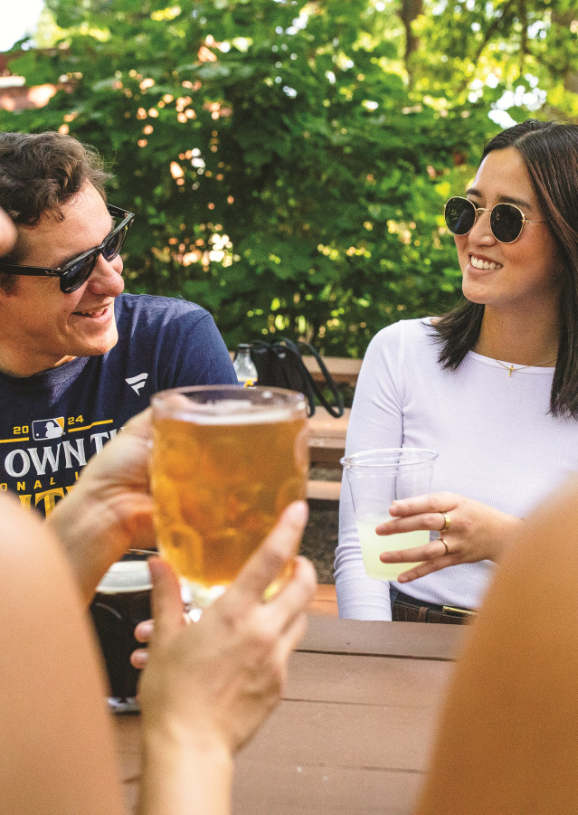 Friends gather around a picnic table at Hubbard Park Beer Garden, chatting and holding drinks beneath leafy trees on a sunny afternoon. Several people wear sunglasses while laughing and talking in the shaded outdoor seating area.