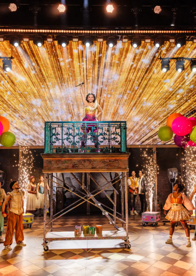 A large ensemble performs onstage at the Marcus Performing Arts Center during a colorful musical number. One performer stands atop a raised balcony platform at center stage holding a microphone, framed by a gold starburst LED backdrop and bright balloon clusters. Sparks shoot upward on both sides of the platform while dancers in vibrant costumes pose across the checkered stage beneath sweeping spotlights.