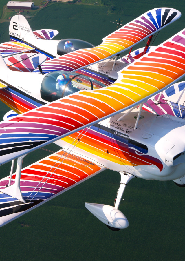 Two brightly colored biplanes fly in close formation above green water, showcasing coordinated aerobatic precision. Their vibrant red, orange, blue, and white striped wings create a striking visual display against the calm background below.
