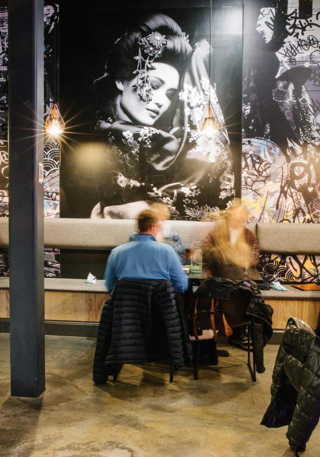 A modern dining space featuring a striking mural of a woman, with patrons seated at tables, enjoying their meals in a vibrant atmosphere.