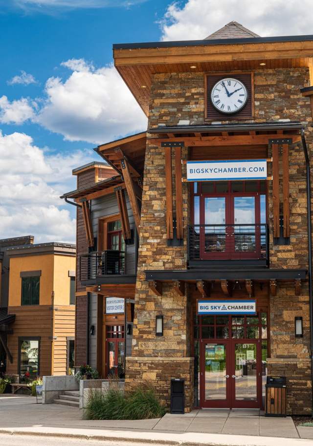 A charming stone building with a clock, featuring the Big Sky Chamber sign. Nearby shops and greenery enhance the inviting streetscape under a blue sky.