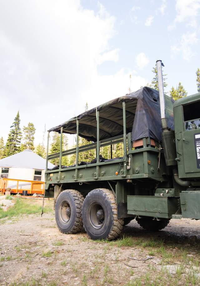 A large vintage army truck with the words 'Montana Yurt Dinner" on the door in front of a cozy yurt with a porch nestled in the trees.