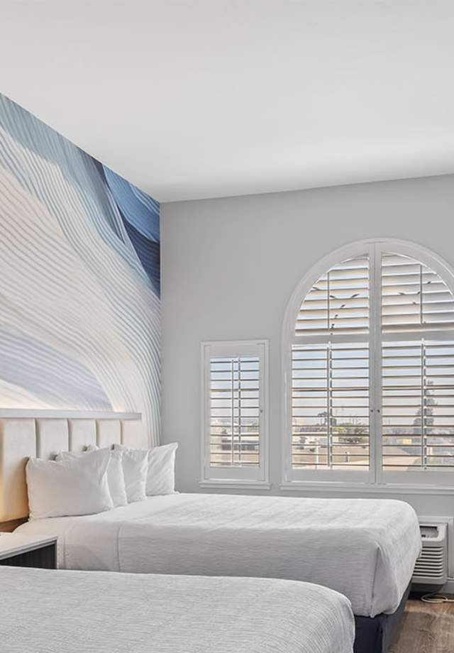 Hotel Skye SFO Double Queen Bedroom. Beds with white bedding in a well-lit room with wood-paneled flooring.