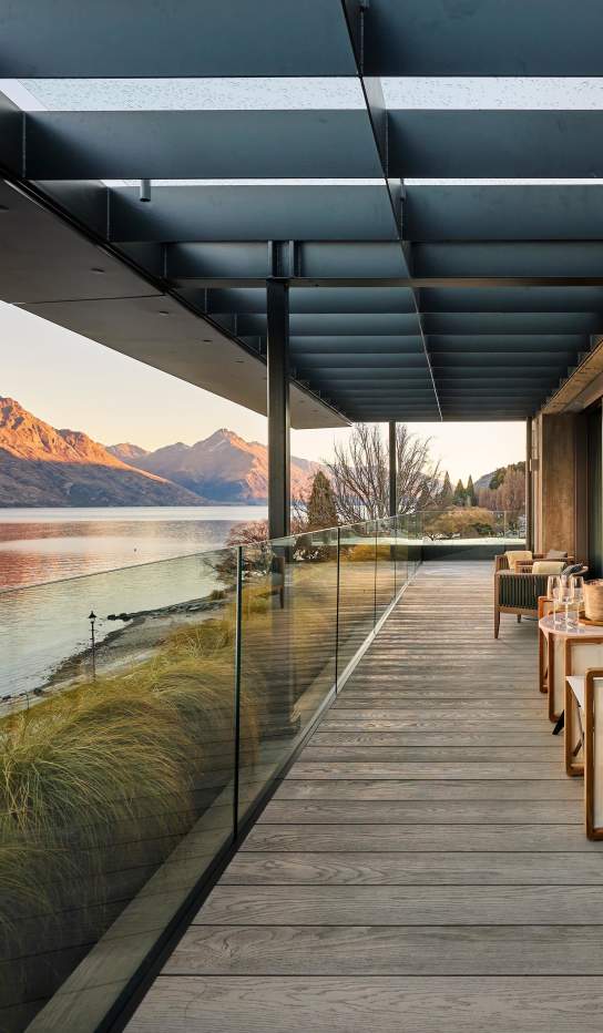 Modern lakeside lodge veranda with glass balustrade overlooking calm water and alpine mountains at sunset, featuring outdoor seating and warm natural timber finishes.