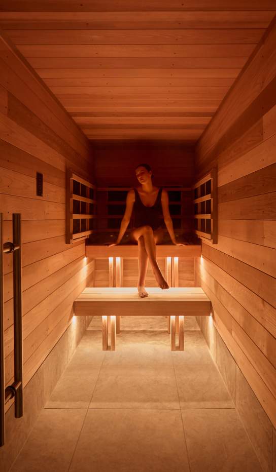 Warm timber-lined sauna interior with soft ambient lighting, featuring a person relaxing on a wooden bench in a modern wellness setting.