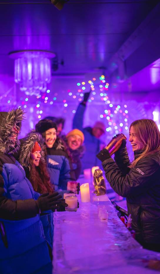 An underground Ice Bar with guests wearing big winter coats standing at a frozen bar with the bar tender making a cocktail.