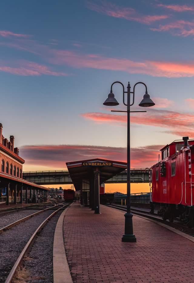 A red train caboose with a holiday wreath sits parked on train tracks in front of a historic train station at sunset.