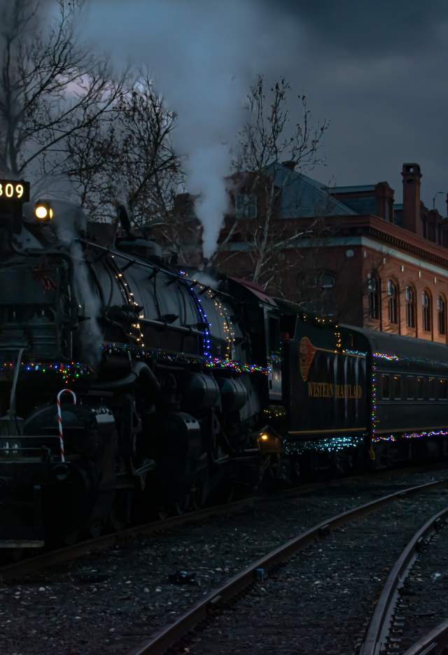A steam engine decorated with Christmas lights and a holiday wreath on the engine is parked in front of a historic brick depot at night.