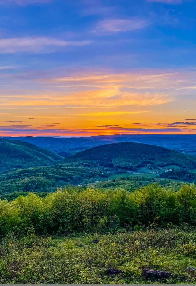The sky is painted in orange, yellow, purple and blues as the sun sets over the mountains along the grassy mountain edge.