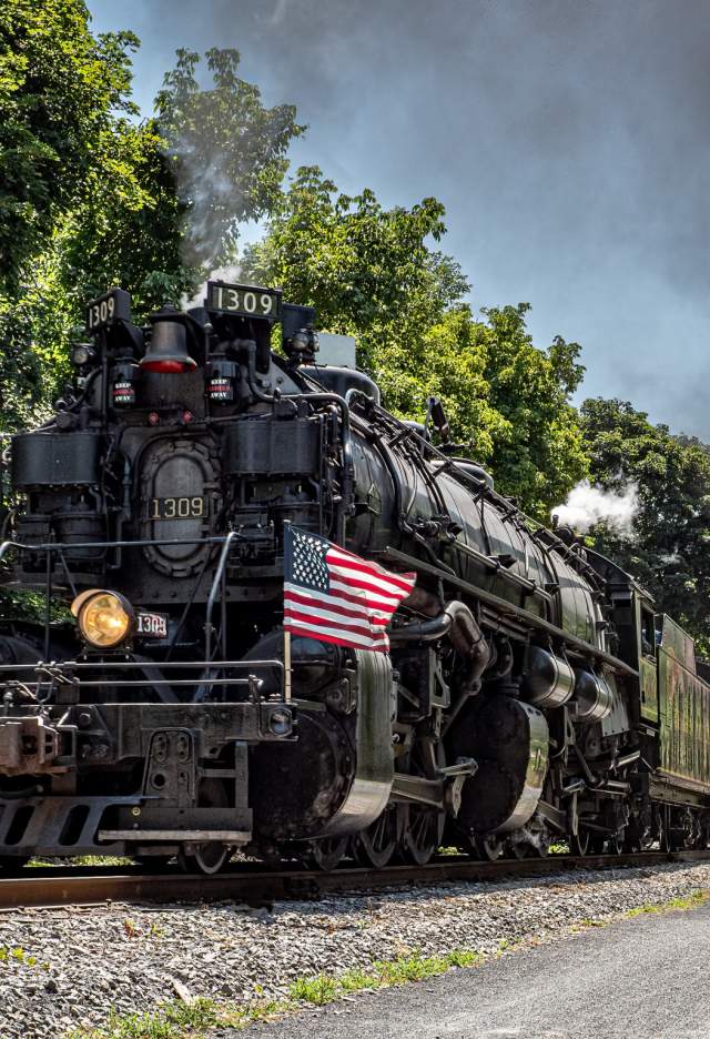 A large black steam engine runs along train track in the summer with smoke billowing from front and two American flags on either side of the engine.