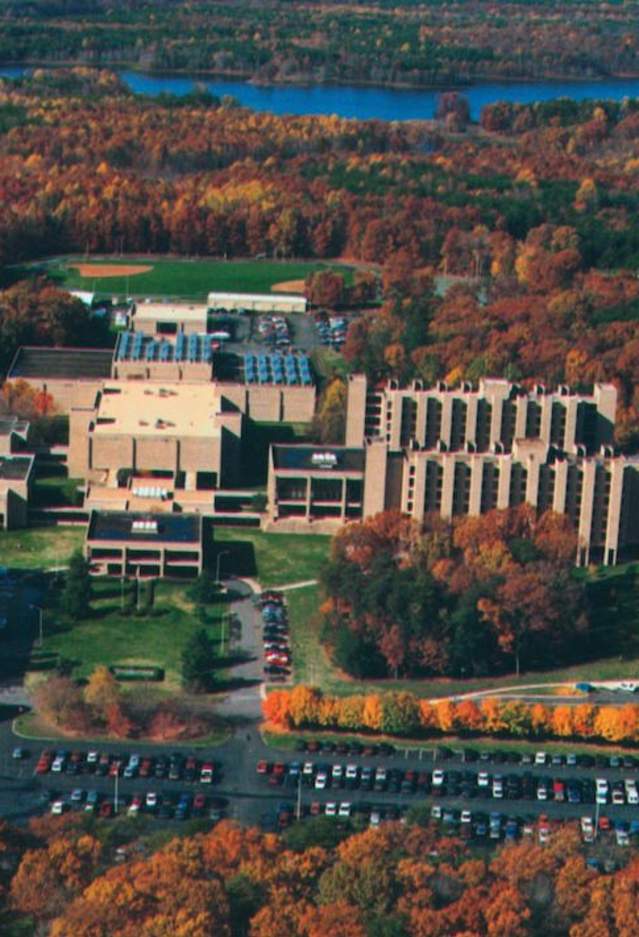 a birds eye view of a campus with buildings, a parking lot filled with cars, surrounded by trees with fall foliage