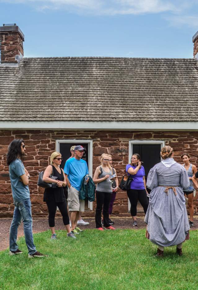 A cobblestone building with visitors and a woman in historic clothing telling a story