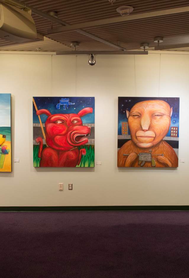 Four paintings on a wall, a yellow dog, a red dog, an abstract brown animal, and a blue dog.