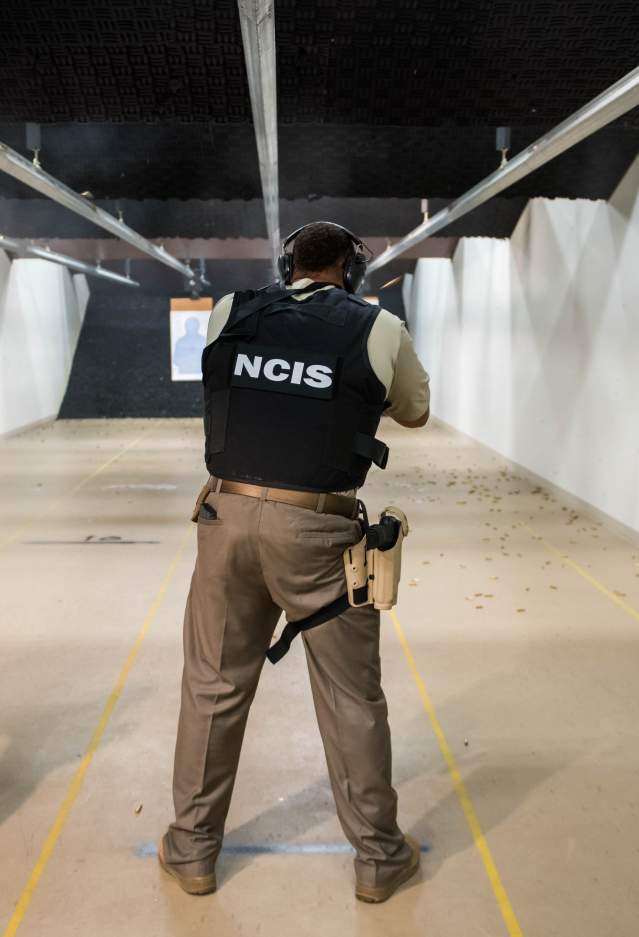 3 men pointing away from the camera, all with black vests on. The 2 men on the left have vests that say "NCIS" and the man on the right has "federal agent" written on the back of his vest. They are holding guns at an indoor gun range.