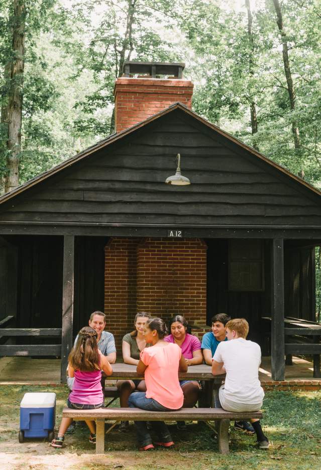 7 people sitting at a picnic bench in front of a cabin in a forest
