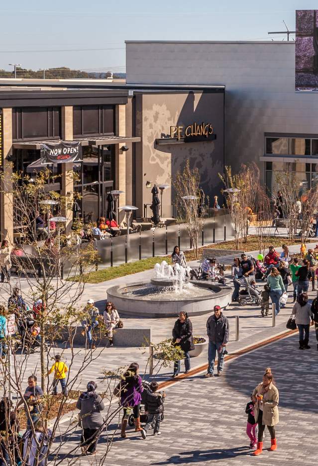 people walking around the courtyard of a shopping area, in the courtyard there is a fountain, patio heaters, benches, trees and tents as well as storefronts.