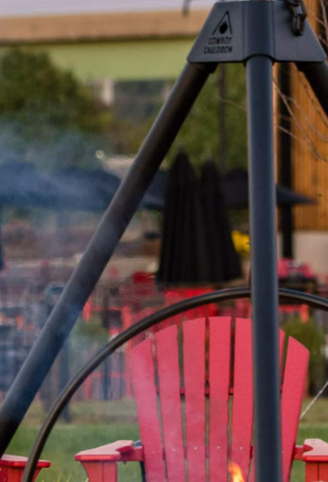 People sitting around a fire in red Adirondack chairs, in the back there is a building and an outdoor stage