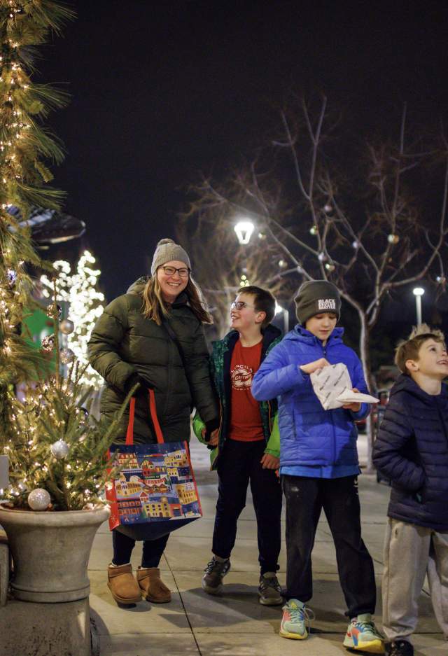A group of two adults and three children walk together on a decorated city sidewalk at night during the holiday season. They are bundled in winter coats, hats, and gloves. Festive lights and small decorated trees line the shop windows on the left. One adult carries a colorful tote bag, and one child holds a snack. Everyone appears cheerful and engaged with one another as they enjoy the winter evening.”