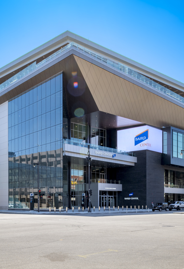 A daytime exterior view of the Baird Center in Milwaukee, showing its modern glass façade, angular roofline, and skywalk connection over the street, with cars and pedestrians nearby.