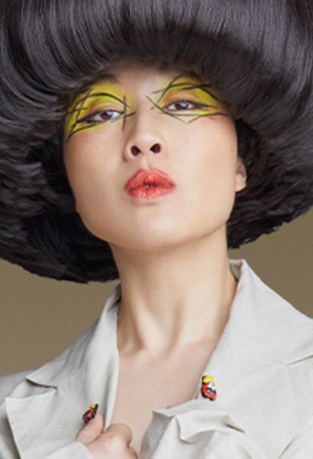 a headshot of Atsuko Okatsuka against a neutral background. They wear a light-colored oversized jacket and striking makeup with bright yellow eyeshadow and red lips, creating a bold, avant-garde look.