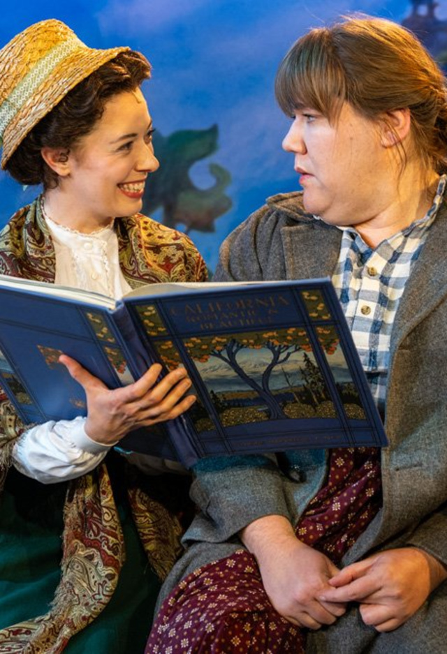 Two actors sit on a stage as one reads from a book and the other reacts with surprise.