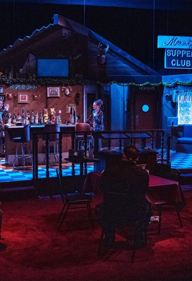 Actors perform on stage in a cozy, dimly lit theater set designed as a small-town supper club, with audience members seated at tables close to the stage for an intimate viewing experience.