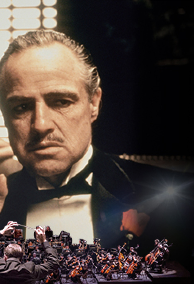 A large screen shows a close-up of two men in tuxedos, with one whispering to the other. In front of the screen, an orchestra plays with a conductor standing at the center, facing the musicians.