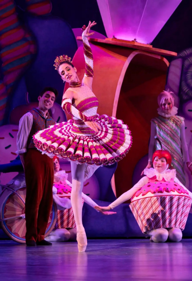 A ballerina in a red-and-white candy-themed costume performs a graceful pose on stage, surrounded by dancers dressed as sweets, including cupcakes and confectioners. The colorful set features oversized candies and whimsical decorations, illuminated by pink and purple stage lighting.