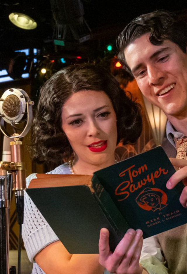 Two actors dressed in 1940s-style clothing perform in a vintage radio studio setting. The woman, with short curled hair, and the man, wearing a vest and tie, smile as they read from a script labeled “Tom” in front of an old-fashioned microphone. A third person stands in the background near festive holiday lights.