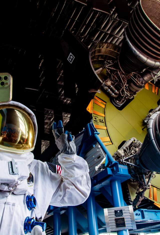A visitor wearing a NASA astronaut suit takes a selfie beneath the massive Saturn V rocket engines inside the U.S. Space & Rocket Center in Huntsville, with the towering rocket display filling the background of the museum exhibit.