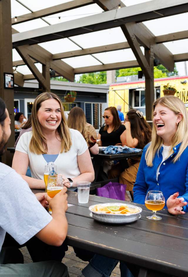 Friends laughing over craft beers and chips with guacamole at a picnic table on the covered patio at Lion's Tail Brewing in Wauwatosa, with a food truck parked in the background.