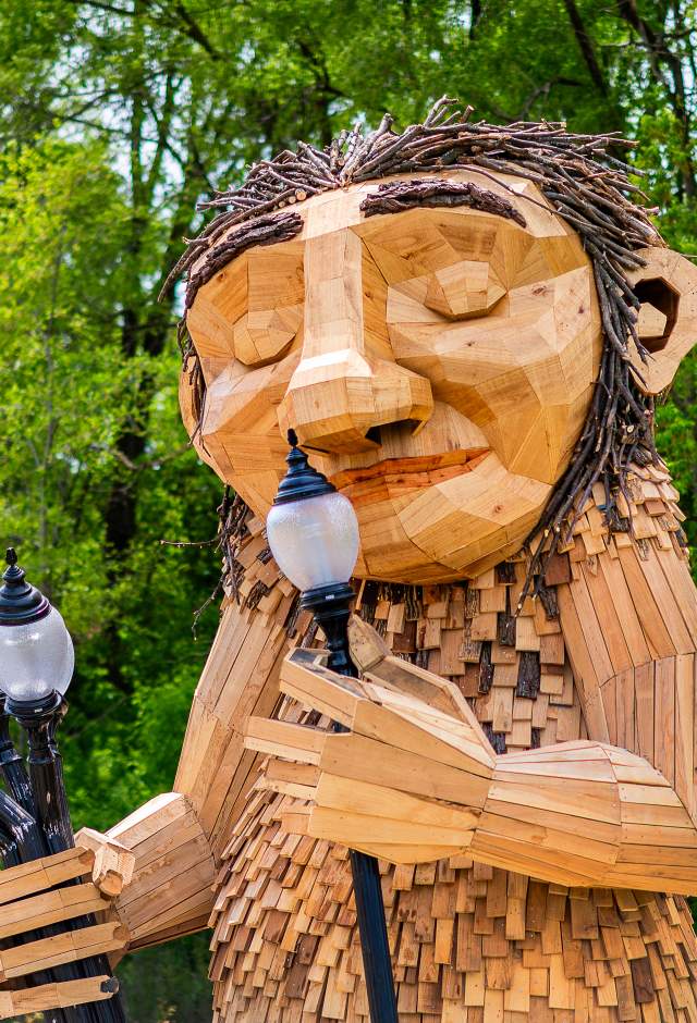 A large wooden sculpture of 'Mama Rosa,' a Thomas Dambo troll installation in Wauwatosa. The friendly giant figure is constructed from reclaimed wood pieces in natural tones, featuring a carved face with a gentle smile and twig-like hair made from bundled branches. The troll holds a bouquet of old-fashioned street lamps in one hand while lifting one lamp to her nose. The sculpture is set against a lush green forest backdrop with dense tree canopy, creating a whimsical scene that blends art with nature.