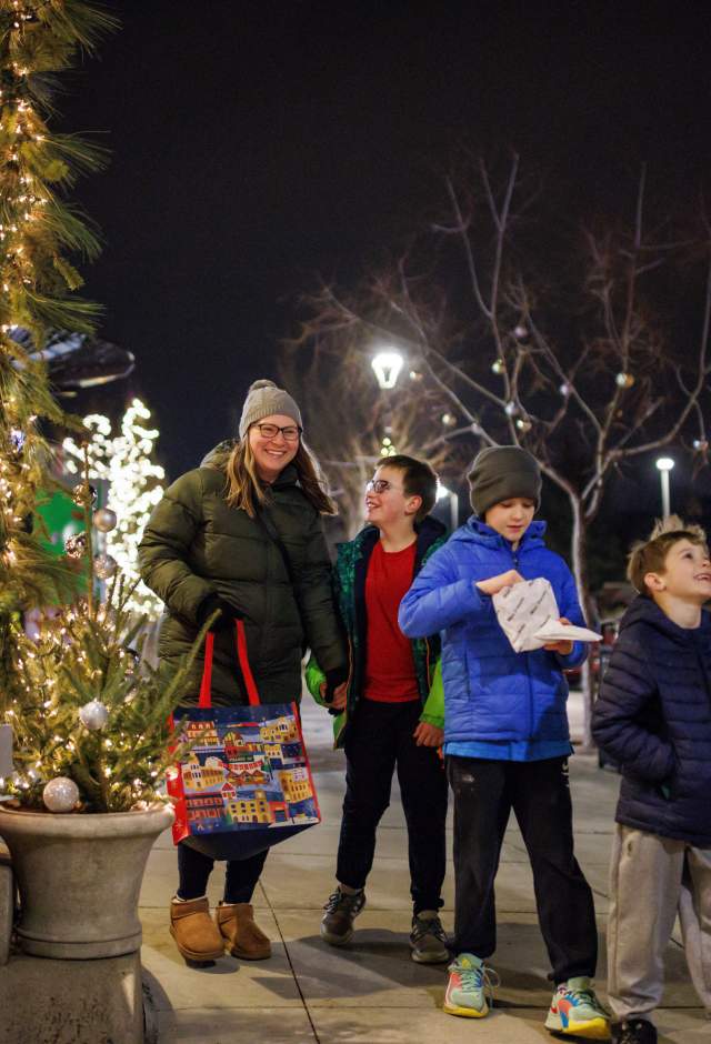 A group of two adults and three children walk together on a decorated city sidewalk at night during the holiday season. They are bundled in winter coats, hats, and gloves. Festive lights and small decorated trees line the shop windows on the left. One adult carries a colorful tote bag, and one child holds a snack. Everyone appears cheerful and engaged with one another as they enjoy the winter evening.”