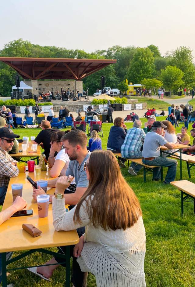 Festivalgoers sit at picnic tables and on lawn chairs across a grassy park while a band performs on an outdoor stage at HartFest.